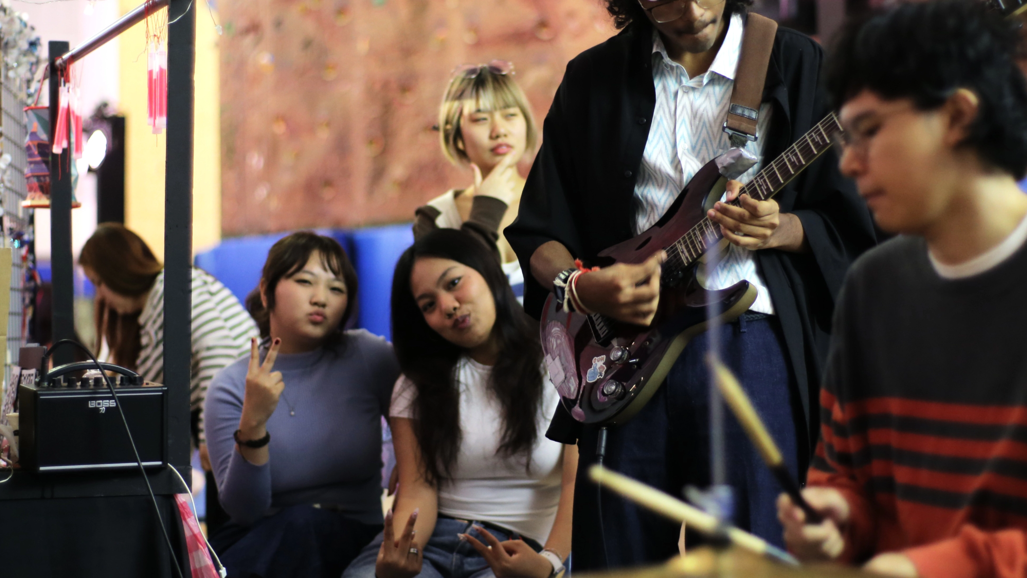 Two girls posing to the cameraman. In front of them are an electric guitarist and a drummer performing.