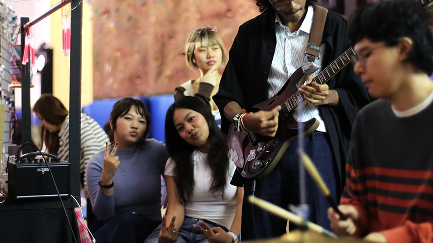 Two girls posing to the cameraman. In front of them are an electric guitarist and a drummer performing.