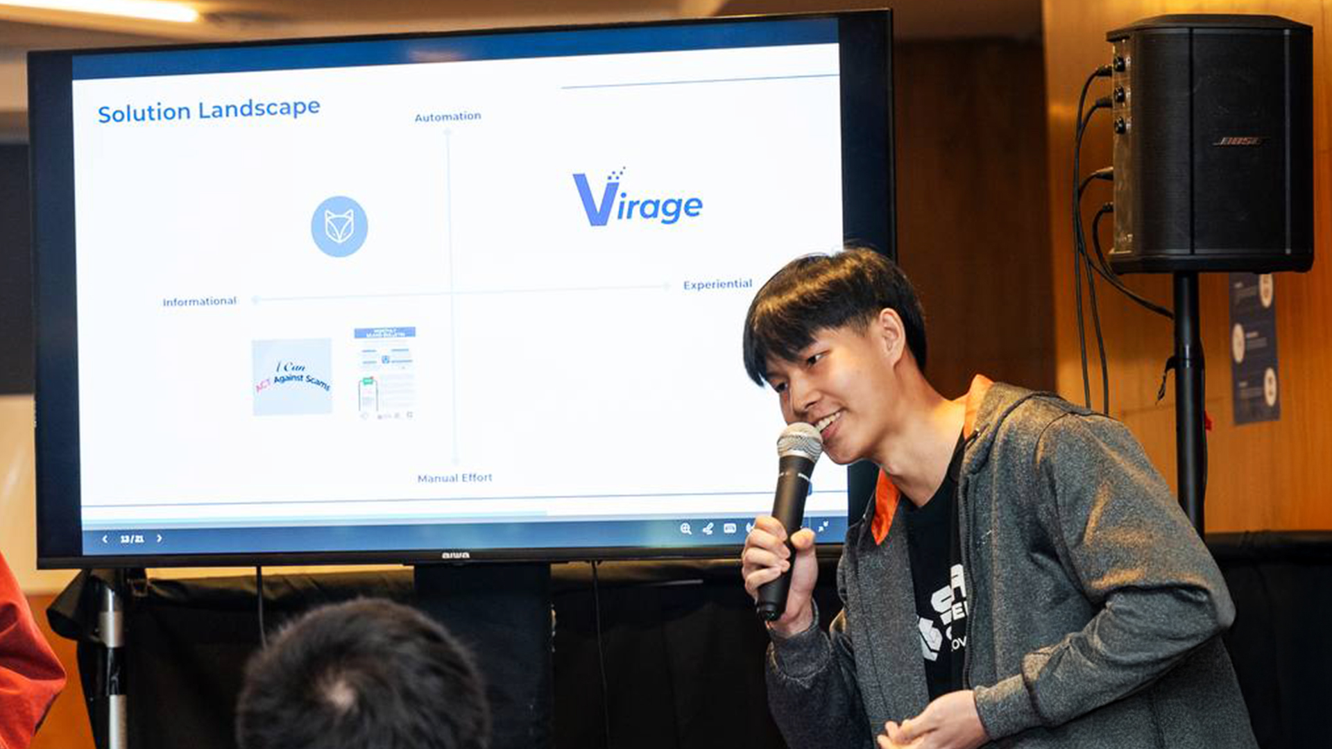 Matthew Wong presenting about Virage; he stands beside a TV screen showing a slide about the solutions that Virage created.
