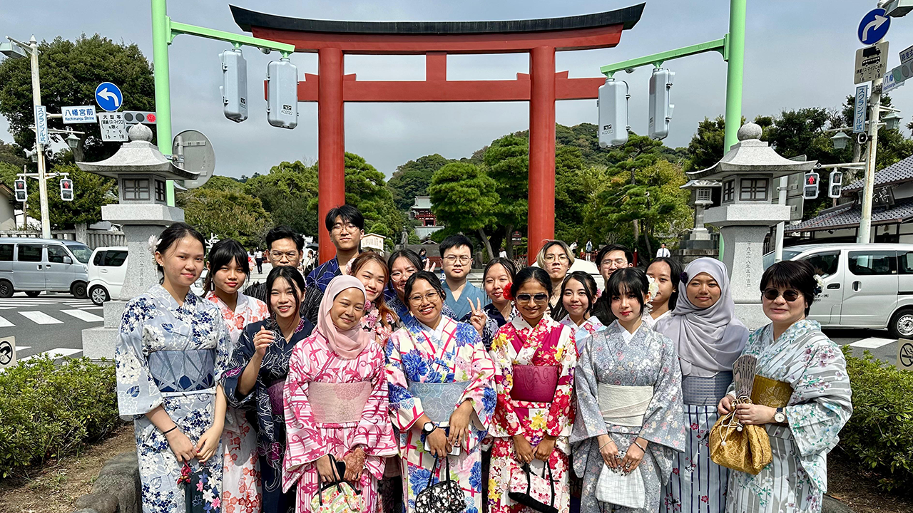 19 students and lecturers in kimonos posing in front of a large red torii gate on a public road.