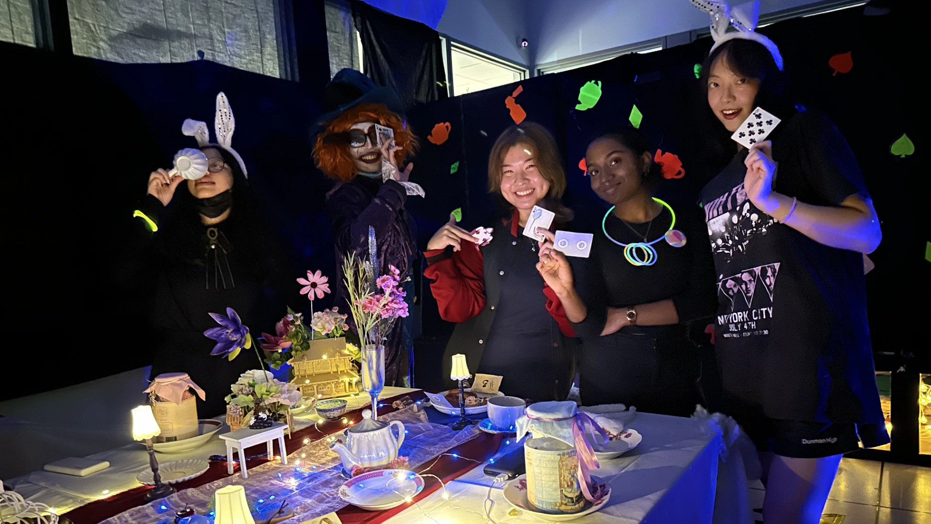 Five students posing at an Alice in Wonderland–themed tea table, holding on to playing cards while wearing bunny ears and a Mad Hatter hat.