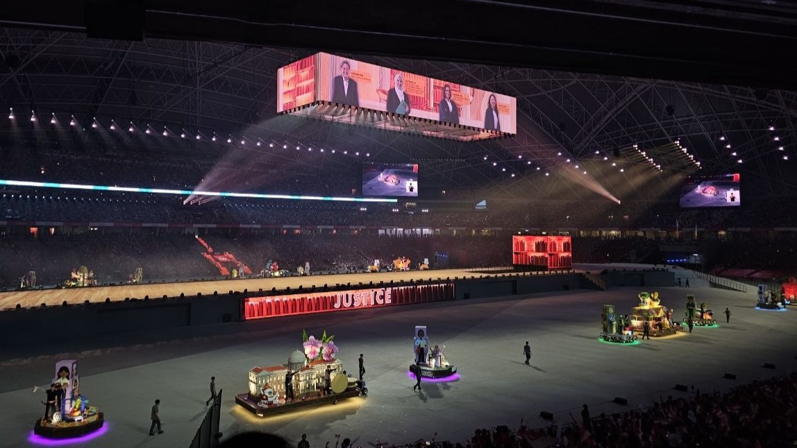 A spectacular view of the NDP 2026 stage setup at the Kallang Sports Hub, featuring the suspended Cuboid screen overhead, the glowing PerfCube on the right of the stage, and the central Fascia banner illuminating the word "Justice."